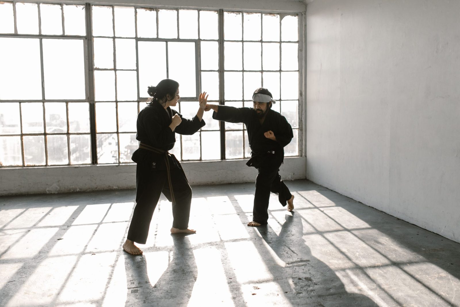 Two martial artists practice barefoot in a sunlit room with glass windows, showcasing skill and focus.